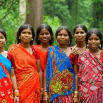 Un ritratto di gruppo di membri di una comunità indigena che vive ai margini della Riserva della Tigre di Mudumalai, India meridionale. Le persone, di età diverse, sono vestite con abiti tradizionali colorati e sorridono. Sullo sfondo, la fitta vegetazione della foresta. Fotografia di ritratto, obiettivo da 35mm, profondità di campo per mettere a fuoco il gruppo ma mantenere visibile il contesto forestale, luce naturale dorata del tardo pomeriggio.