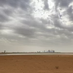 Immagine panoramica della regione desertica di Al Qassim in Arabia Saudita, scattata con un obiettivo grandangolare da 10mm, che mostra un cielo drammatico con un mix di sole e nuvole minacciose che suggeriscono il cambiamento climatico. Il focus è nitido sul terreno arido, con lo skyline di una città futuristica che si intravede nella foschia in lontananza. Lunga esposizione per rendere le nuvole setose e l'acqua, se presente, liscia.
