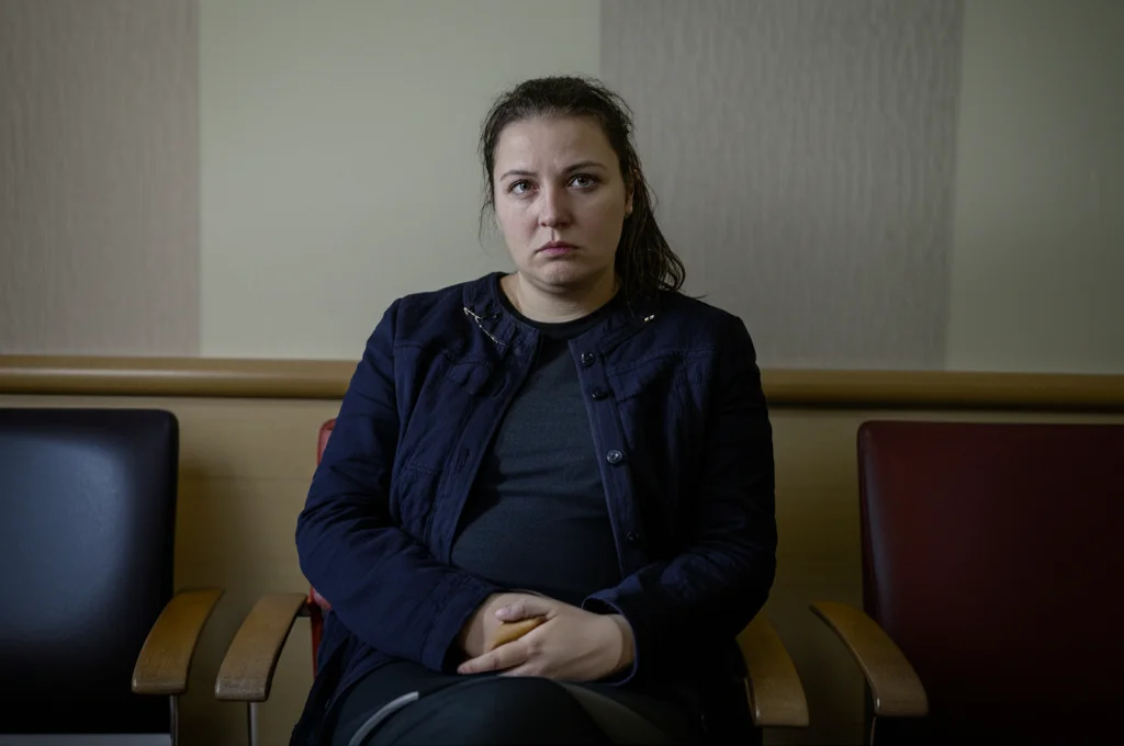 Una donna Nera incinta, con un'espressione pensierosa e preoccupata, siede in una sala d'attesa di un ospedale. La luce è soffusa, quasi cupa. Fotografia ritratto, obiettivo 35mm, toni bicromatici blu e grigio per accentuare la serietà, profondità di campo che sfoca leggermente lo sfondo per concentrarsi sull'emozione della donna.