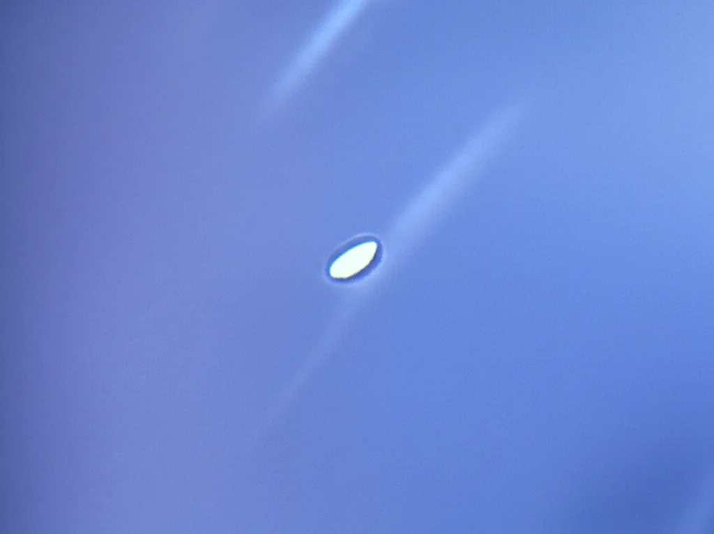 Macro fotografia di un guanto chirurgico latex-free teso, con una luce che evidenzia una micro-perforazione quasi invisibile. Obiettivo macro 100mm, alta definizione, illuminazione controllata per enfatizzare la texture e il difetto del guanto.