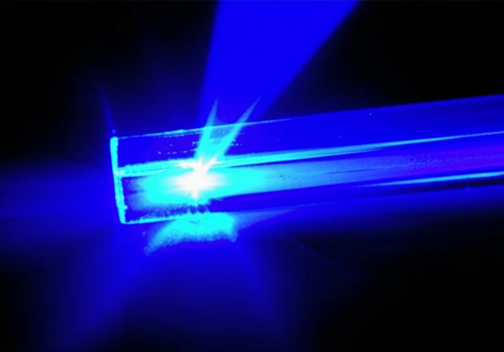 Macro fotografia, obiettivo 90mm, di una cuvetta di quarzo contenente una soluzione limpida di dutasteride in acetonitrile, posizionata all'interno di uno spettrofluorimetro. Un fascio di luce UV (non visibile direttamente ma implicato) la colpisce, e la soluzione emette una debole fluorescenza bluastra. Illuminazione da laboratorio controllata, alta definizione, messa a fuoco precisa sulla cuvetta e sulla luce emessa, sfondo scuro per esaltare la fluorescenza.