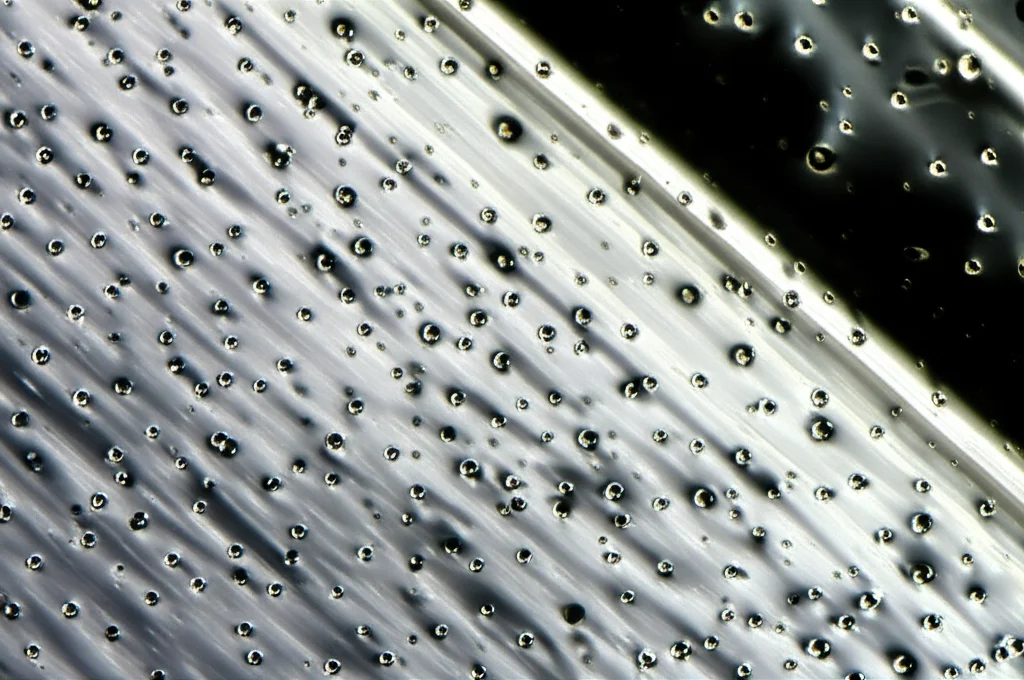 Macro fotografia di nanoparticelle metalliche sospese in un fluido trasparente, che scorre su una superficie metallica porosa e riscaldata. Illuminazione controllata per evidenziare la texture della superficie e il movimento del fluido. Obiettivo macro 100mm, alta definizione, focus preciso sulle particelle e sulla superficie.