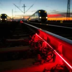 Immagine fotorealistica di un raggio laser rosso debole che viaggia all'interno di un cavo in fibra ottica trasparente e scintillante, posato accanto a un binario ferroviario ad alta velocità al crepuscolo, obiettivo grandangolare 18mm, lunga esposizione per catturare il movimento sfocato di un treno AV sullo sfondo e la traccia luminosa del laser, messa a fuoco nitida sulla fibra e sul binario vicino.