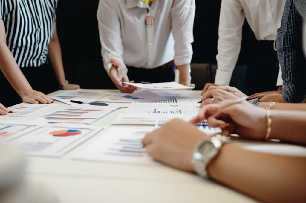 Fotografia di un team multidisciplinare (infermieri, assistenti sociali, ricercatori) durante una riunione di lavoro in un ufficio moderno. Stanno discutendo animatamente attorno a un tavolo pieno di grafici e documenti. Lenti zoom 24-70mm per catturare l'interazione del gruppo, illuminazione controllata da ufficio, focus sull'interazione e sui materiali di lavoro.