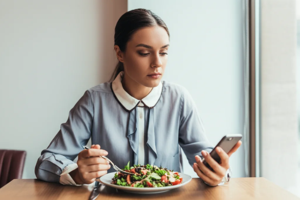 Una donna di circa 30 anni, seduta a un tavolo con davanti un'insalata colorata e il suo smartphone. L'espressione è pensierosa, quasi ansiosa, mentre confronta il suo pasto con qualcosa sullo schermo. Luce naturale da una finestra laterale, effetto leggermente desaturato. Obiettivo da 35mm, profondità di campo che sfoca leggermente lo sfondo.