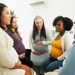 Fotografia stile ritratto di un gruppo eterogeneo di donne incinte, culturalmente diverse, che partecipano attivamente a una discussione sulla ricerca sanitaria prenatale in un ambiente luminoso e accogliente. Obiettivo prime 35mm, luce naturale diffusa, profondità di campo accentuata per isolare leggermente il gruppo dallo sfondo, espressioni coinvolte e collaborative, toni caldi.