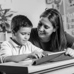 Un'aula scolastica luminosa e accogliente, un bambino autistico di circa 8 anni è seduto a un banco e lavora concentrato su un tablet con un'insegnante di sostegno al suo fianco che lo guida con un sorriso. L'ambiente è sereno e inclusivo, con materiali didattici colorati sullo sfondo. Fotografia stile ritratto, obiettivo 35mm, luce naturale diffusa, profondità di campo che sfoca leggermente lo sfondo per mettere a fuoco il bambino e l'insegnante, bianco e nero con elevato contrasto, stile film noir.