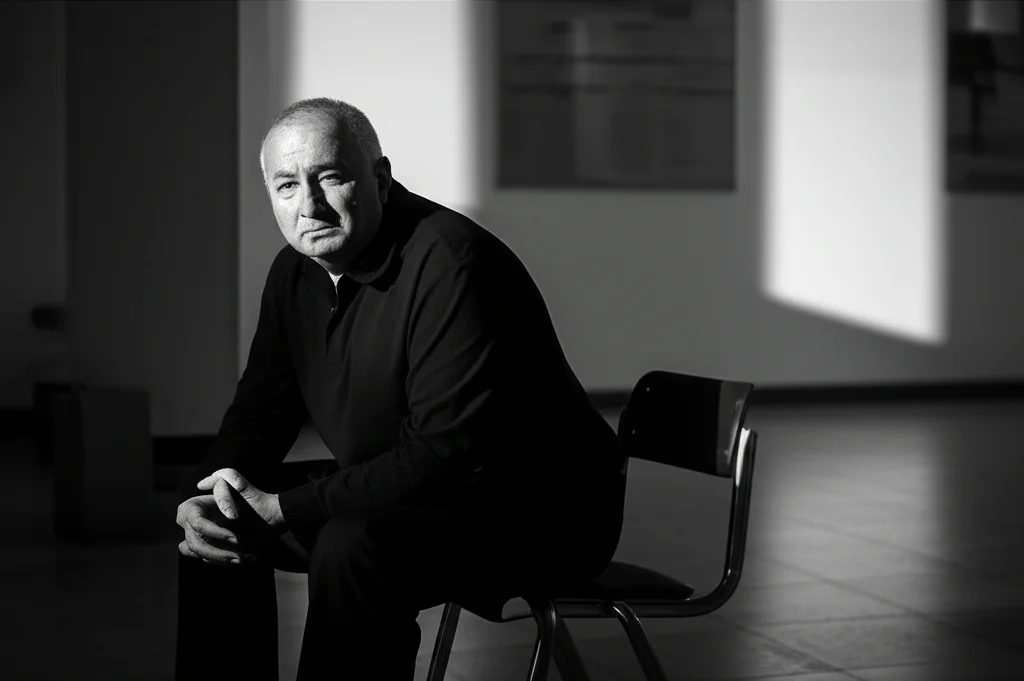 Un ritratto toccante di un familiare, un uomo di mezza età, seduto in una sala d'attesa di un ospedale, lo sguardo perso nel vuoto. Fotografia in bianco e nero, obiettivo da 35mm, profondità di campo che sfoca leggermente lo sfondo, mettendo in risalto l'espressione pensierosa e preoccupata. Stile film noir per accentuare la tensione emotiva.