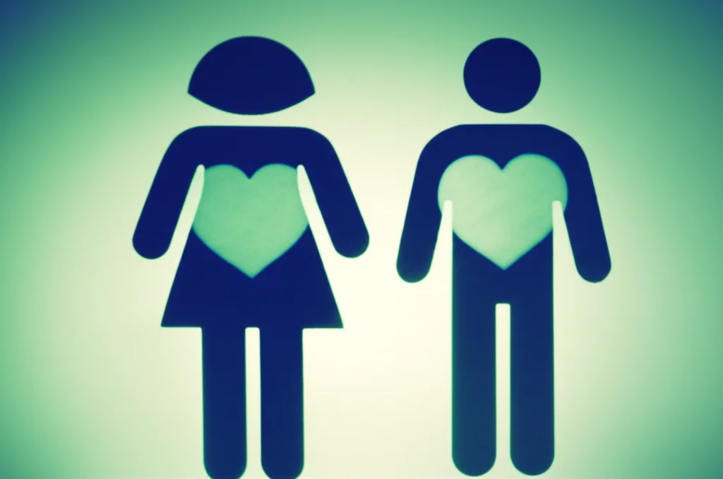 Due figure stilizzate, una maschile e una femminile, con accanto un cuore luminoso. La figura femminile ha un cuore leggermente più grande e luminoso, per simboleggiare la maggiore protezione cardiovascolare. Lo scatto è realizzato con un obiettivo da 35mm, utilizzando un effetto duotone blu e verde acqua, con una profondità di campo che mette a fuoco le figure e i cuori, lasciando lo sfondo leggermente sfocato.