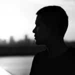 Fotografia di ritratto, un uomo migrante interno a Shenzhen, Cina, in un contesto urbano moderno ma affollato, guarda pensieroso verso l'orizzonte al tramonto, simboleggiando la riflessione sulla salute, il futuro e la prevenzione della tubercolosi latente, obiettivo 35mm, bianco e nero, profondità di campo.