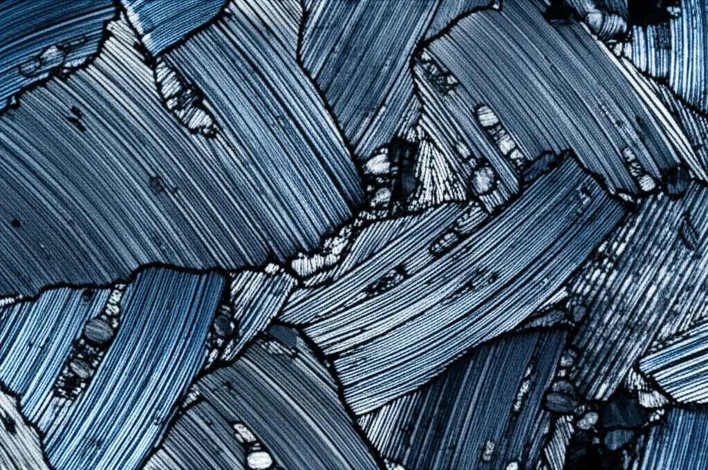 Immagine fotorealistica di un campione di acciaio AHSS al microscopio elettronico, che mostra chiaramente le diverse fasi microstrutturali come ferrite, bainite e martensite. Illuminazione laterale per evidenziare la tridimensionalità delle fasi. Obiettivo macro 100mm, alta definizione, colori leggermente desaturati con accenti blu per un look scientifico.