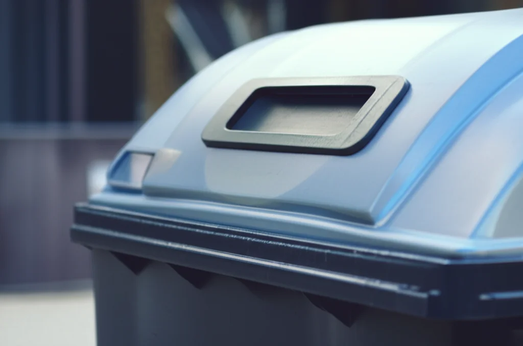 Un cassonetto intelligente in un contesto urbano moderno, ripreso con obiettivo macro da 85mm per evidenziare il sensore di livello e i rifiuti al suo interno, alta definizione, illuminazione controllata per un look pulito e tecnologico, con un leggero effetto bokeh sullo sfondo per concentrare l'attenzione sul cassonetto.