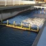 Visualizzazione del flusso d'acqua complesso attorno a piloni di ponte gemelli in un canale a U sperimentale, con palette sommerse visibili a monte. Obiettivo 35mm, stile fotorealistico, profondità di campo che evidenzia l'interazione tra strutture e corrente.