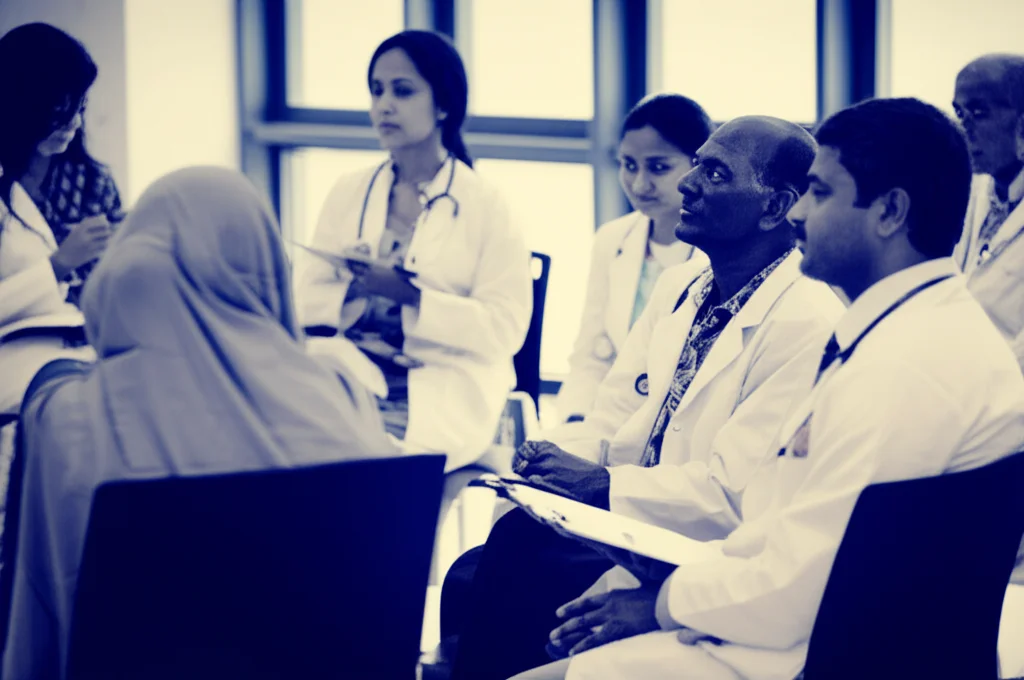 Un gruppo eterogeneo di pazienti indiani con diabete di tipo 2, di età diverse, che partecipano a una sessione informativa sullo studio clinico in un moderno centro medico in India. Alcuni ascoltano attentamente, altri prendono appunti. Luce naturale soffusa. Obiettivo prime da 35mm, profondità di campo per mettere a fuoco il gruppo ma sfocare leggermente lo sfondo. Tonalità seppia e blu per un effetto filmico.