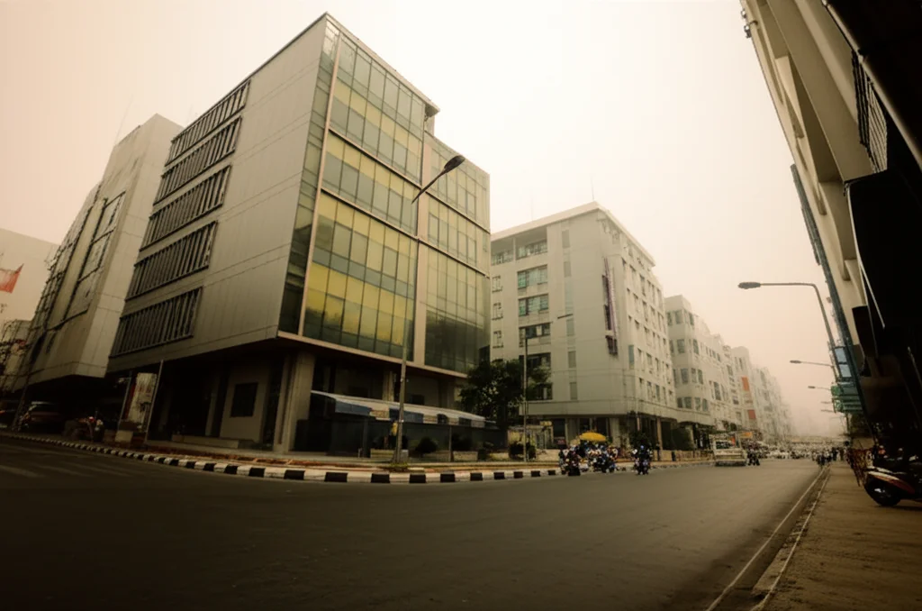 Veduta a livello stradale di un'area urbana densamente popolata nel distretto di Bangkoknoi, con un mix di edifici residenziali moderni e tradizionali. Si percepisce un leggero smog o foschia. Alcuni motorini parcheggiati. Wide-angle lens, 24mm, focus nitido sull'ambiente urbano.