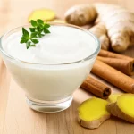 Yogurt bianco cremoso in una ciotola di vetro trasparente, guarnito con un rametto di timo fresco. Accanto alla ciotola, su una superficie di legno chiaro, sono disposte elegantemente alcune stecche di cannella e pezzi di radice di zenzero fresco. Luce naturale laterale controllata che esalta la texture dello yogurt. Obiettivo macro 90mm, alta definizione, messa a fuoco precisa sullo yogurt e sulle spezie in primo piano, sfondo leggermente sfocato.
