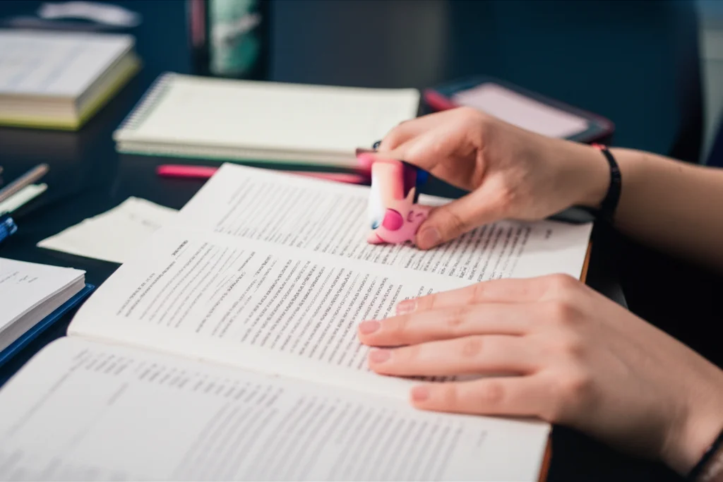 Primo piano di mani di studenti che indicano un libro di testo medico aperto su un tavolo, con altri materiali di studio sparsi intorno. Illuminazione controllata, obiettivo macro 100mm, alta definizione dei dettagli del testo e della carta, colori vivaci.