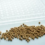 Fotografia macro ad alto dettaglio di granuli di mangime per animali su una superficie pulita, con una piastra ELISA a 96 pozzetti parzialmente visibile e sfocata sullo sfondo. Illuminazione controllata laterale per creare texture e profondità, obiettivo macro 90mm, messa a fuoco selettiva sui granuli.