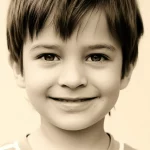 Ritratto fotografico di un bambino piccolo, circa 3 anni, con pelle visibilmente migliorata dopo un trattamento per dermatite atopica. Il bambino sorride serenamente. Luce naturale e morbida, obiettivo da ritratto 35mm, profondità di campo per mettere a fuoco il viso e la pelle sana. Duotone seppia e crema per un tocco delicato.