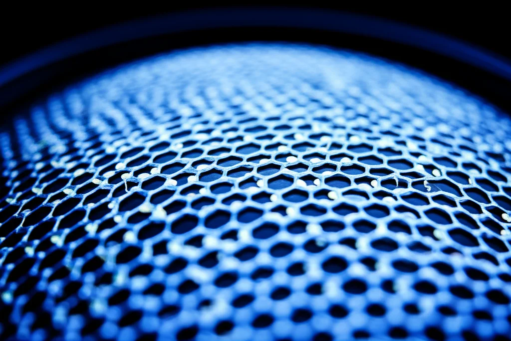 Macro fotografia di un sottile strato di materiale nanostrutturato, come il grafene, illuminato per evidenziare la sua texture. Si vedono percorsi luminosi astratti che rappresentano i fononi che si muovono attraverso il materiale, alcuni dei quali collidono tra loro. Lente macro 100mm, high detail, precise focusing, controlled lighting con una leggera dominante bluastra per dare un'idea di tecnologia avanzata.