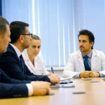 Fotografia realistica e professionale di un gruppo eterogeneo di docenti universitari di odontoiatria in una sala riunioni luminosa, impegnati in una discussione seria ma costruttiva sui metodi di valutazione degli studenti. Obiettivo zoom 24-70mm impostato a 35mm, luce morbida da finestra, focus nitido sui volti espressivi.