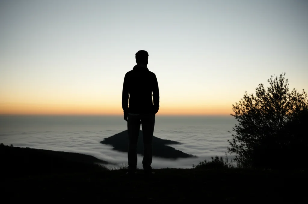Silhouette di una persona in piedi su una collina che guarda un paesaggio vasto e nebbioso all'alba, simboleggiando la complessità, l'incertezza ma anche la speranza nella gestione di una condizione cronica come la fibromialgia con comorbidità psicologiche. Fotografia paesaggistica, obiettivo grandangolare 24mm, luce soffusa e atmosferica, colori tenui.