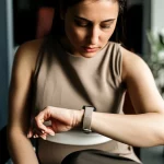 Immagine fotorealistica di una donna che guarda pensierosa il suo smartwatch al polso, simbolo del monitoraggio dei sintomi dell'endometriosi e della fatica. Obiettivo 50mm prime lens, profondità di campo ridotta per focalizzare l'attenzione sulla donna e l'orologio, luce naturale morbida che entra da una finestra.