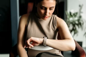 Immagine fotorealistica di una donna che guarda pensierosa il suo smartwatch al polso, simbolo del monitoraggio dei sintomi dell'endometriosi e della fatica. Obiettivo 50mm prime lens, profondità di campo ridotta per focalizzare l'attenzione sulla donna e l'orologio, luce naturale morbida che entra da una finestra.