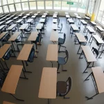 Foto grandangolare di un'aula scolastica moderna e luminosa vista dall'alto, con banchi vuoti disposti ordinatamente. Luce naturale che entra dalle finestre, messa a fuoco nitida su tutta la scena, obiettivo 14mm, simboleggia l'ambiente scolastico oggetto di studio per interventi sul benessere emotivo.