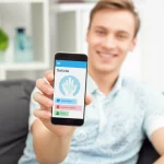 Fotografia realistica in primo piano di uno smartphone che mostra un'app per la salute mentale con grafici e messaggi di supporto, tenuto da mani giovanili. Sullo sfondo sfocato, un giovane sorridente e rilassato in un ambiente luminoso e confortevole. Obiettivo prime 50mm, profondità di campo ridotta, luce naturale morbida, colori vivaci ma non saturi.