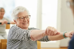 Persona anziana sorridente che esegue un leggero stretching su una sedia durante una sessione di gruppo post-ictus. Fotografia ritratto, obiettivo 50mm, luce morbida da finestra, sfondo leggermente sfocato, colori caldi.