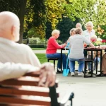Un gruppo diversificato di anziani sorridenti che partecipano insieme a un'attività ricreativa all'aperto, come giardinaggio o una partita a carte in un parco. Luce solare calda, colori vivaci. In contrasto, in un angolo dell'immagine, leggermente sfocato, un anziano osserva da solo, seduto su una panchina, con un'espressione neutra o leggermente malinconica. Obiettivo 35mm per catturare la scena ampia ma permettere il dettaglio emotivo. L'immagine simboleggia il contrasto tra connessione sociale e solitudine/stigma.