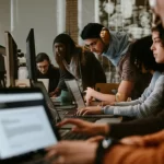 A diverse group of computer science students working collaboratively on laptops in a modern university library, some looking at screens displaying code or text documents, hinting at the integration of technology in their writing process, 35mm portrait lens, depth of field.