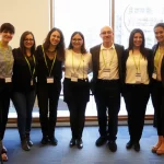 Gruppo multietnico di persone sorridenti che collaborano durante un incontro informativo sulla salute a Bergen, Norvegia, simboleggiando gli ambasciatori di salute migranti. Fotografia di ritratto di gruppo, obiettivo prime 35mm, luce naturale brillante da una finestra, profondità di campo che mette a fuoco il gruppo ma lascia intravedere l'ambiente di una sala riunioni accogliente.