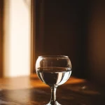 Immagine simbolica di un bicchiere mezzo pieno e uno mezzo vuoto affiancati su un tavolo di legno rustico, con una luce calda e soffusa proveniente da una finestra laterale che illumina maggiormente il bicchiere mezzo pieno, fotografia still life, obiettivo 50mm prime, messa a fuoco selettiva sul bordo del bicchiere pieno, sfondo leggermente sfocato, concetto di ottimismo e prospettiva.
