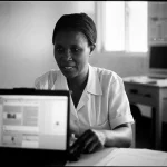 Primo piano di un operatore sanitario del Lesotho, donna, sui 40 anni, con un sorriso rassicurante, mentre utilizza un computer portatile in un ambulatorio rurale ben illuminato. Sullo schermo del portatile si intravede l'interfaccia di un sistema di cartelle cliniche elettroniche. Obiettivo prime 35mm, black and white film, profondità di campo per sfocare leggermente lo sfondo e concentrare l'attenzione sull'operatrice e il computer, trasmettendo professionalità e speranza.