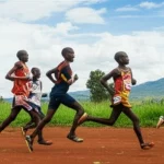 Gruppo di atleti di resistenza ugandesi, maschi e femmine adolescenti, che si allenano insieme correndo all'alba sulle colline ondulate della regione di Sebei, Uganda. Fotografia panoramica con obiettivo grandangolare 18mm, lunga esposizione per rendere le nuvole soffuse nel cielo dorato, messa a fuoco nitida sul paesaggio e sugli atleti in movimento dinamico.