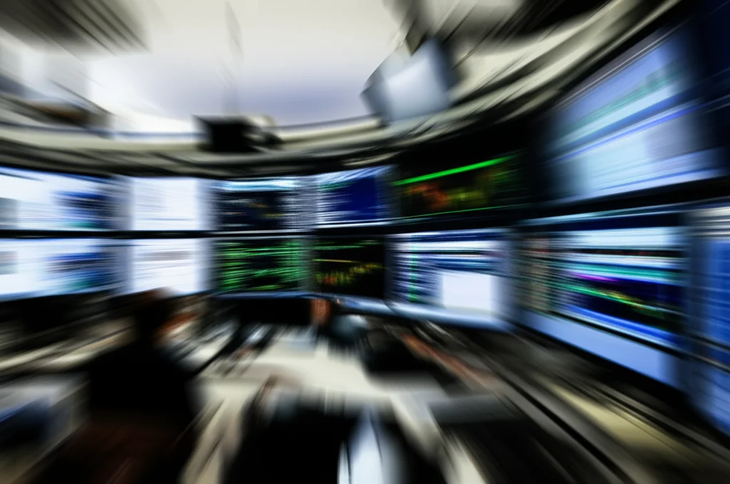 Foto dinamica (teleobiettivo 100-400mm, alta velocità otturatore, tracciamento movimento) che cattura il blur di numeri e grafici su schermi di trading multipli, suggerendo l'intensa attività e volatilità del mercato Bitcoin durante i periodi di segnale dei pattern grafici. L'effetto movimento trasmette urgenza e rapidità decisionale.