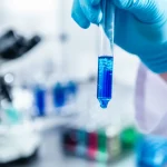Immagine fotorealistica, scattata con lente prime 35mm, che mostra le mani guantate di uno scienziato mentre tengono una provetta in cui un liquido blu vibrante sta visibilmente schiarendo dal basso verso l'alto. Sullo sfondo sfocato si intravede attrezzatura da laboratorio. La profondità di campo mette a fuoco la provetta e il processo di decolorazione enzimatica in corso.