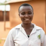 A photorealistic 35mm portrait of a community health worker in Togo, wearing simple clothes, standing in a rural village setting, with a friendly expression, depth of field blurring the background.