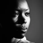 Fotografia di ritratto, una donna dell'Africa Sub-Sahariana con espressione pensierosa e forte, illuminata lateralmente. Stile film noir, bianco e nero ad alto contrasto, obiettivo 35mm, profondità di campo ridotta per focalizzare l'attenzione sul volto e sull'emozione complessa legata alle difficili scelte di salute materna e alle pratiche culturali come le MGF/C.