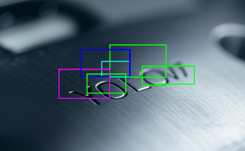 Immagine fotorealistica di un'interfaccia software su schermo che mostra il modello YOLOv7 in azione. Si vedono bounding box colorati che circondano con precisione i caratteri incisi al laser su un'immagine di superficie metallica. Effetto profondità di campo, focus sui box. Tonalità duotone blu e grigio.