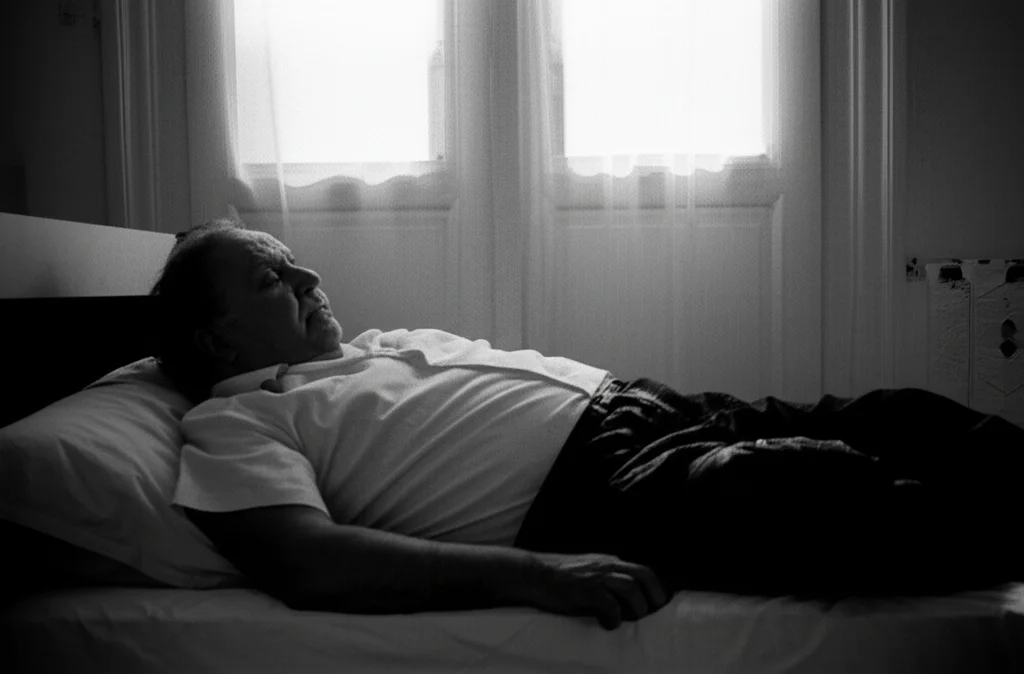 Primo piano di un uomo di mezza età addormentato su un cuscino, leggermente in sovrappeso, con luce soffusa dalla finestra laterale. Fotografia ritratto, obiettivo 35mm, profondità di campo ridotta per sfocare lo sfondo della camera da letto, bianco e nero.