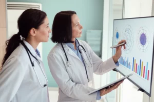 Fotografia di ritratto, due medici di base, un uomo e una donna di mezza età, in un moderno ambulatorio medico, discutono davanti a uno schermo che mostra grafici di indicatori di qualità sanitaria. Obiettivo prime 35mm, profondità di campo, illuminazione controllata, toni neutri.
