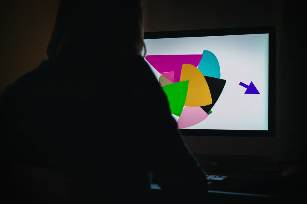 Scena fotorealistica vista da dietro le spalle di una persona seduta davanti a un monitor. Sullo schermo si vedono forme geometriche colorate e una freccia lampeggiante in un angolo. L'ambiente è leggermente scuro per enfatizzare lo schermo. Obiettivo prime 35mm, profondità di campo media, effetto cinematografico con leggera vignettatura.