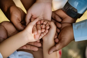 Mani di diversa etnia che si stringono delicatamente attorno alla mano di un bambino piccolo, simboleggiando supporto e comunità. Fotografia di ritratto, obiettivo 50mm, profondità di campo, toni caldi e morbidi, luce naturale.