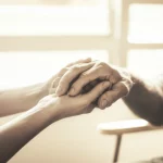 Fotografia di ritratto, obiettivo 35mm, mano di un'infermiera/infermiere che tiene delicatamente la mano di un paziente anziano di etnia diversa, ambientazione serena in una stanza di ospedale o hospice, luce calda e morbida, profondità di campo, colori tenui duotone (es. seppia e crema), espressione di empatia e fiducia.
