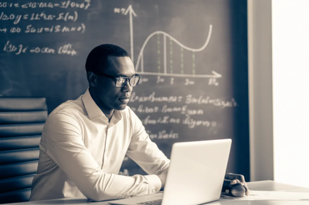 Ritratto di un economista africano, uomo di mezza età con occhiali, che analizza dati su un laptop in un ufficio moderno. Alle sue spalle, una lavagna con grafici e formule macroeconomiche. Fotografia di persone, obiettivo prime 35mm, profondità di campo media, illuminazione naturale da finestra laterale, colori duotone seppia e blu.