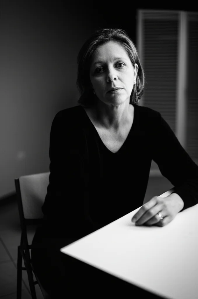 Ritratto fotografico 35mm, in bianco e nero stile film noir, di una madre con espressione preoccupata e stanca seduta a un tavolo spoglio di fronte a una sedia vuota in un'aula scolastica poco illuminata, profondità di campo ridotta che sfoca lo sfondo, simboleggiando la frustrazione, l'isolamento e la lotta nelle interazioni scolastiche per il Piano 504.