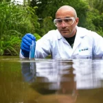 A scientist collecting a water sample from a river in Brazil for eDNA testing, with a focus on the water and surrounding environment. Wide-angle lens, 24mm, sharp focus.